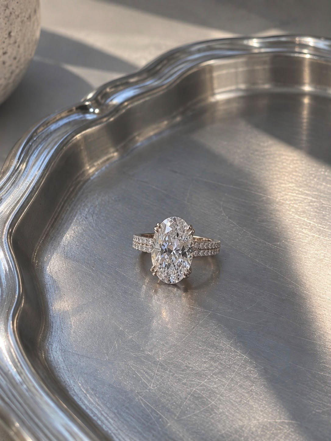 DRITË 3ct oval diamond ring with a double-row pavé band on a scalloped silver tray in sunlight