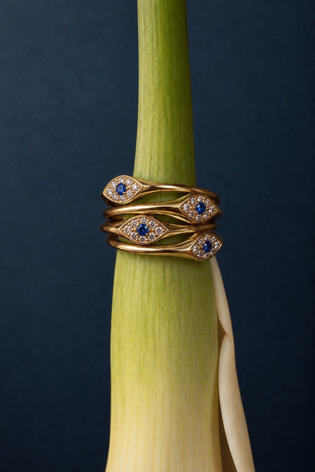 Four FLORA Evil Eye rings stacked on a green stem against a dark petrol backdrop