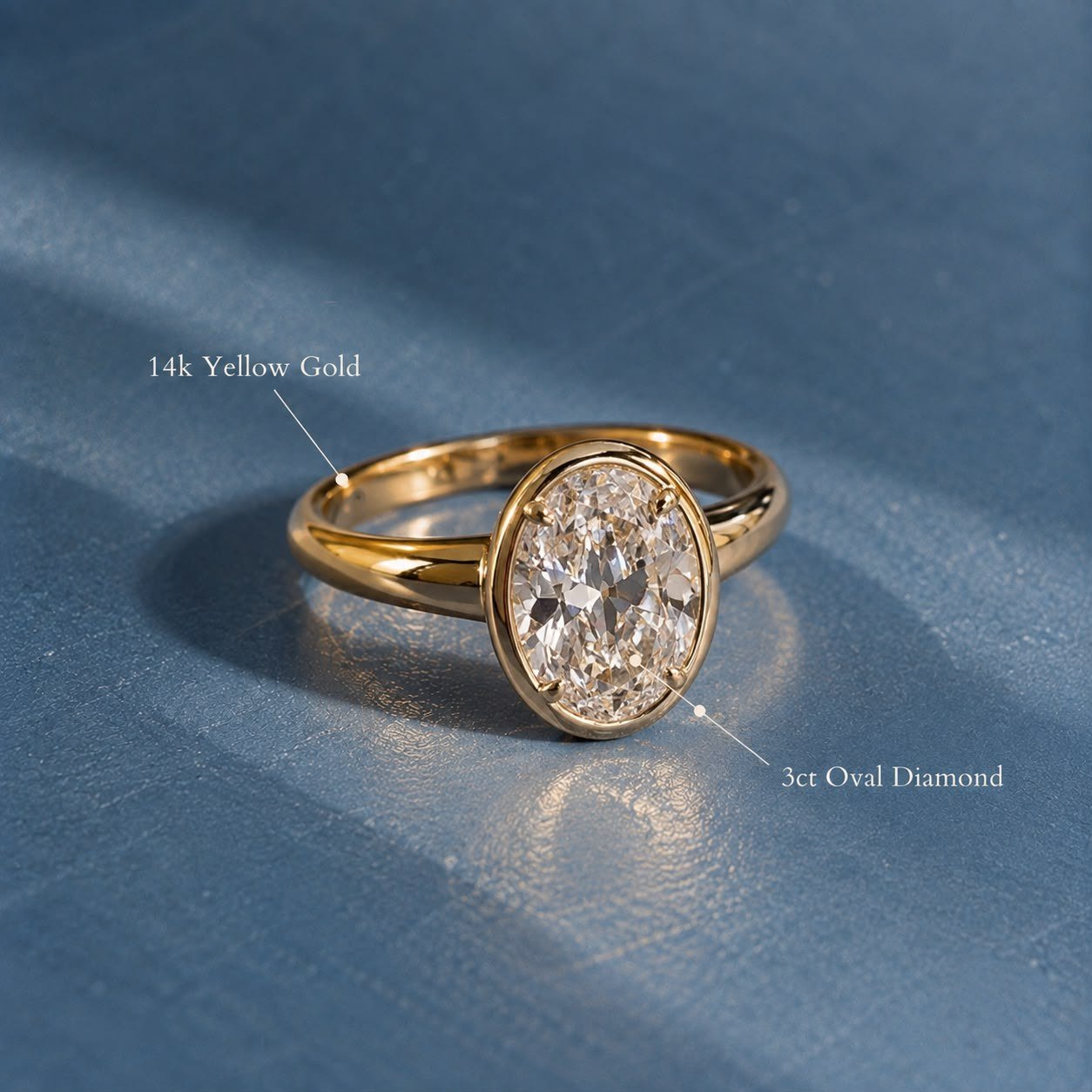 3ct oval lab-grown diamond in a smooth 14K yellow gold bezel on blue linen