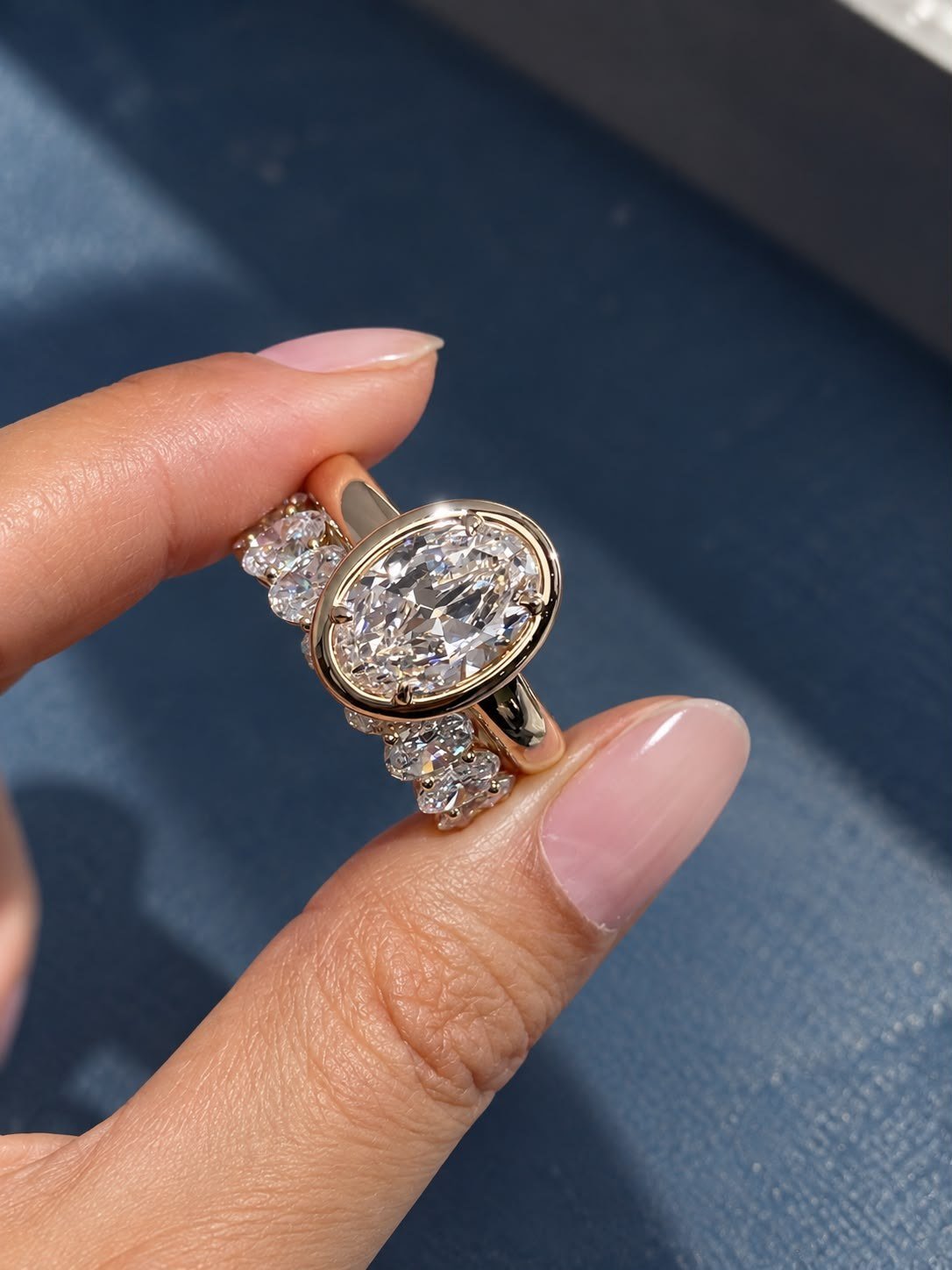 Oval bezel ring held between two fingers in sunlight, beside an oval diamond eternity band