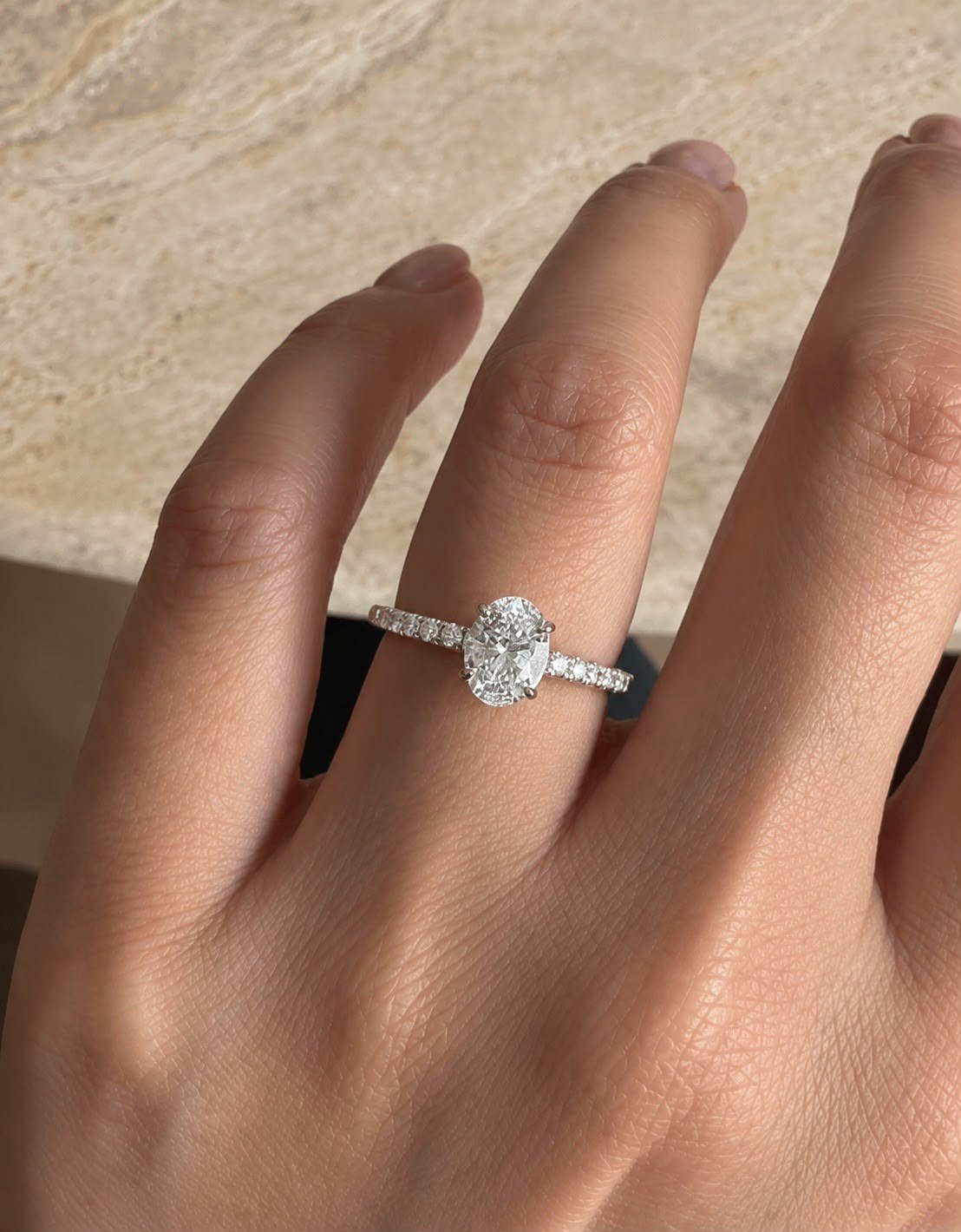 White gold oval pavé engagement ring worn on a hand against travertine