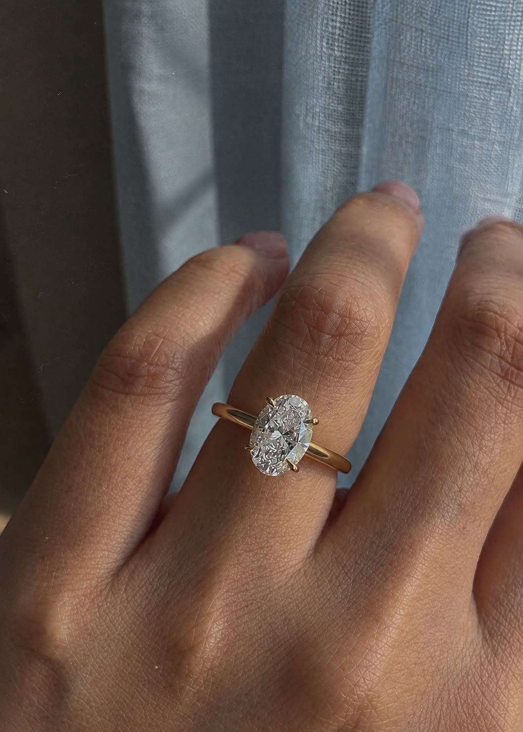 Yellow gold oval solitaire worn on a hand against a pale blue linen curtain